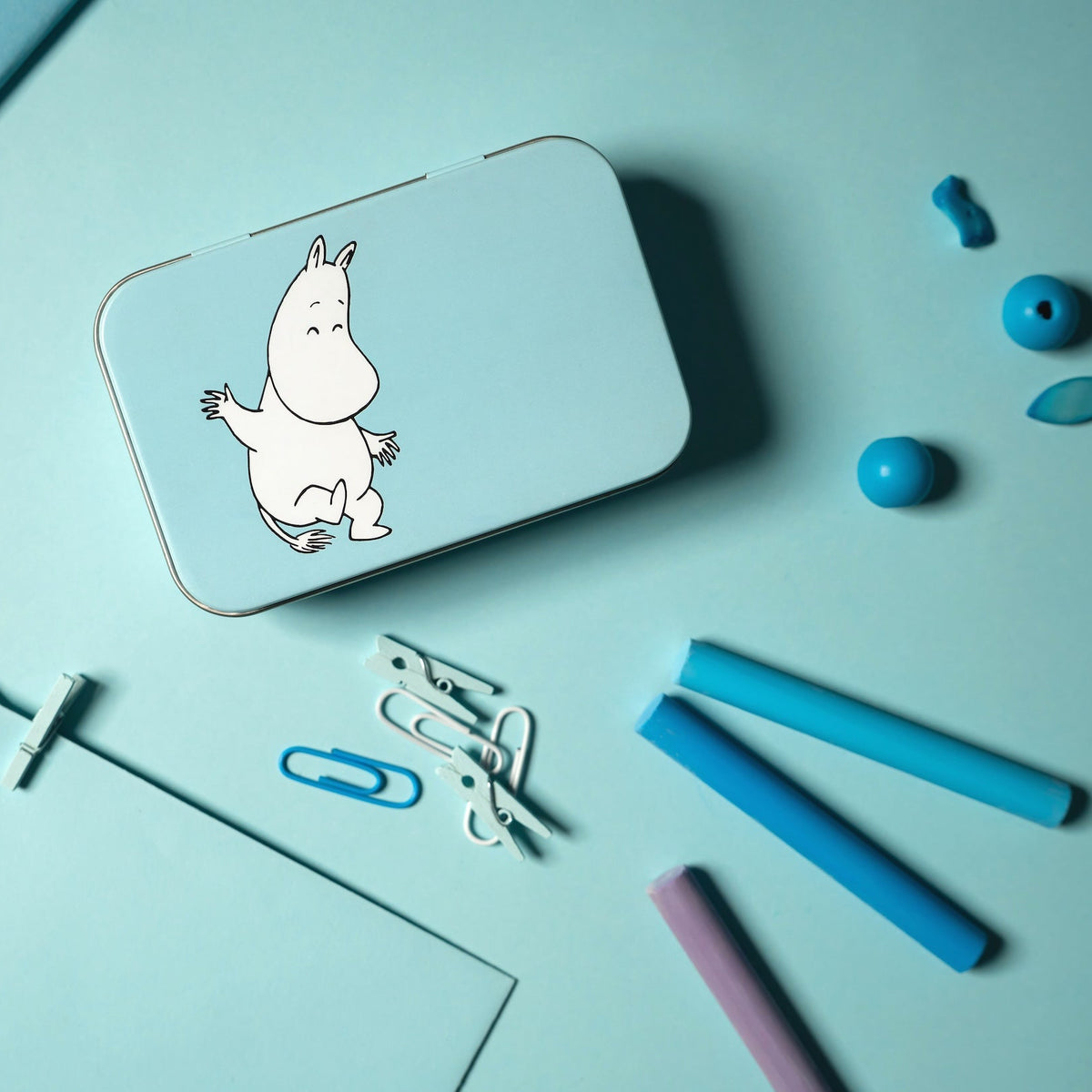 Moomin tin with a Moomintroll on a light blue surface with stationery items.