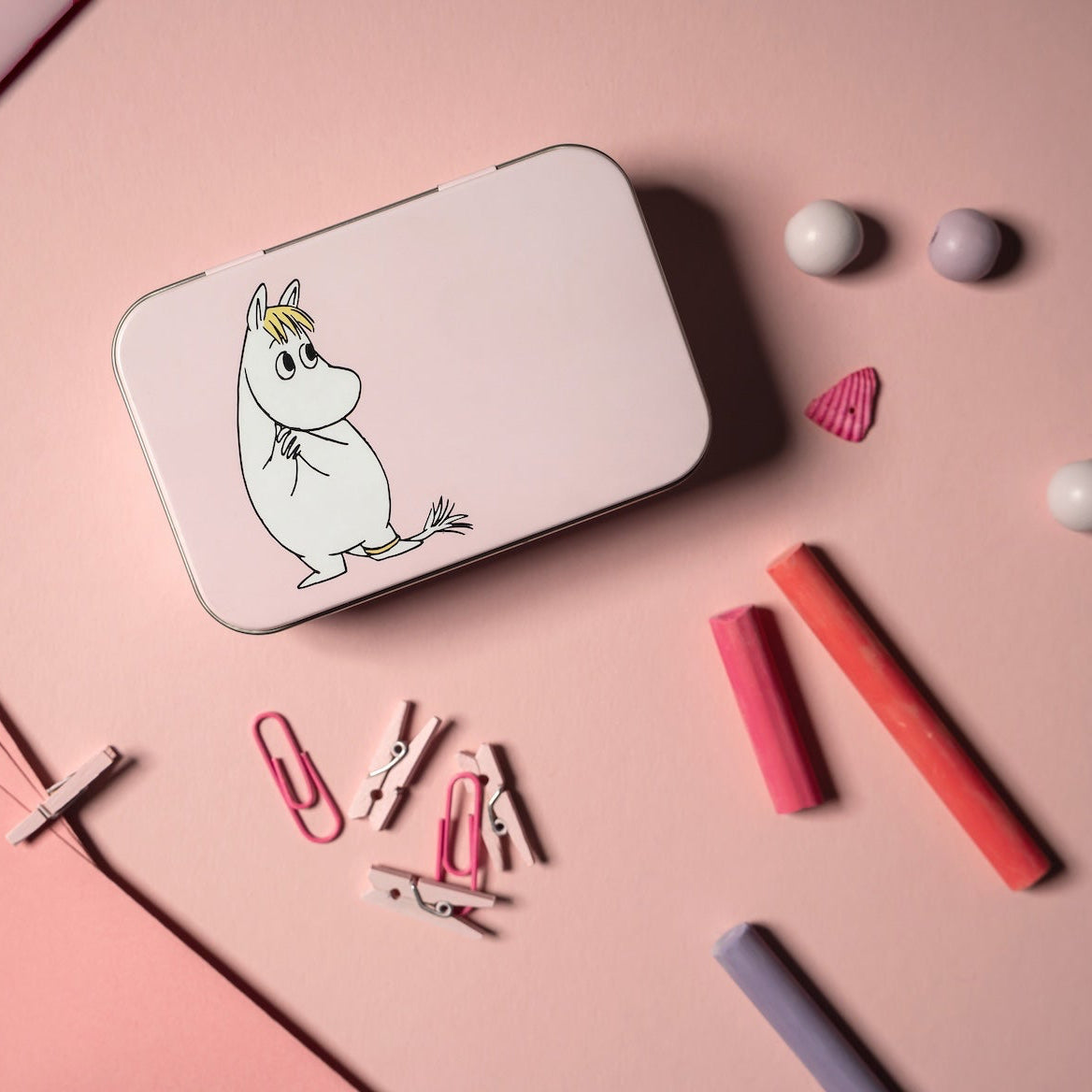 Small rectangular Moomin Snorkmaiden tin on a pink surface with stationery items.