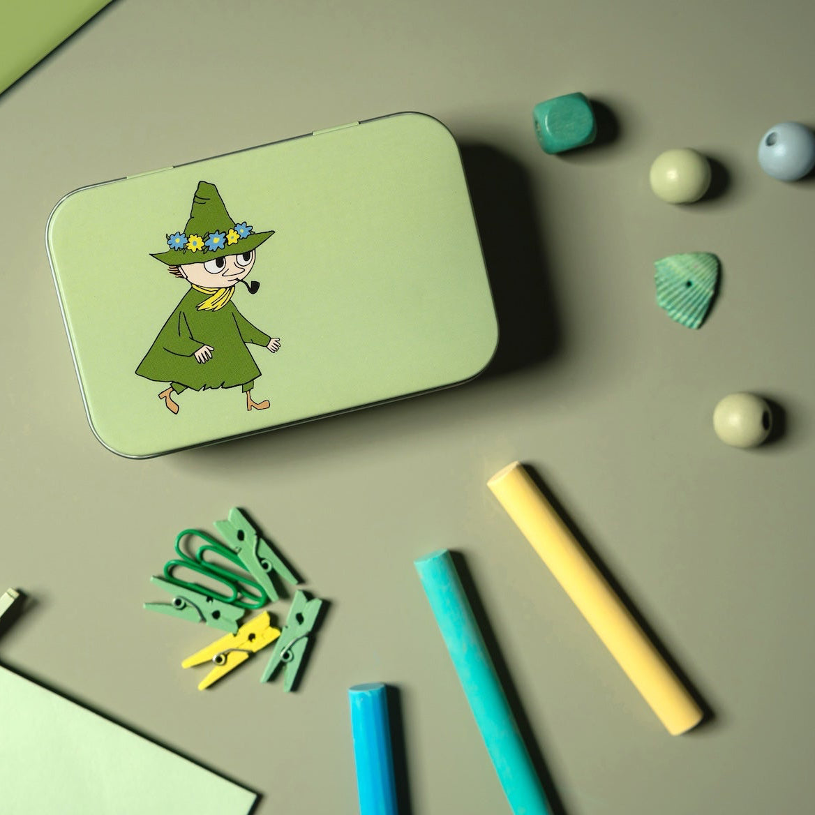 Green tin with Moomin Snufkin surrounded by stationery items on a green surface
