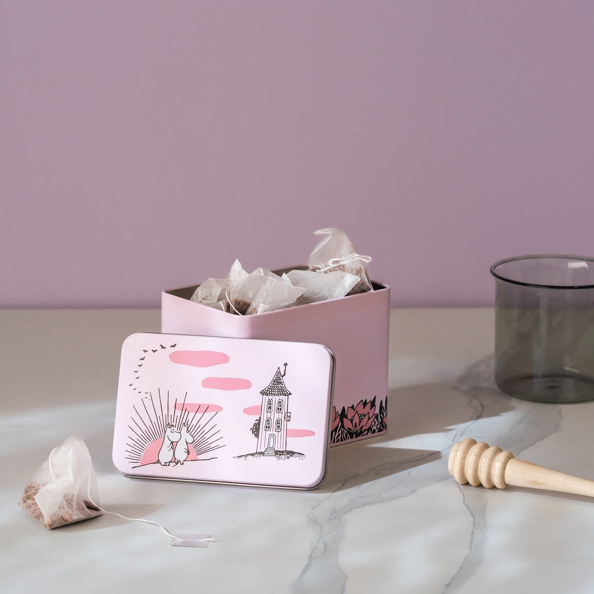 A pink metal Moomin tea tin with a whimsical illustration of a mouse and a house, shown open with tea bags inside on a light marble surface against a mauve background.