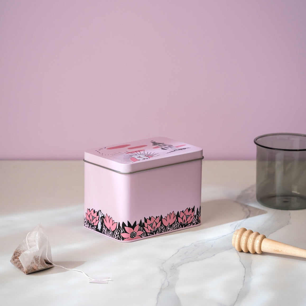 A pink Moomin tea tin with a floral patterned border sits on a white marble surface against a dusty pink background. A teabag, a wooden honey dipper, and a glass cup are arranged around it as styled props.