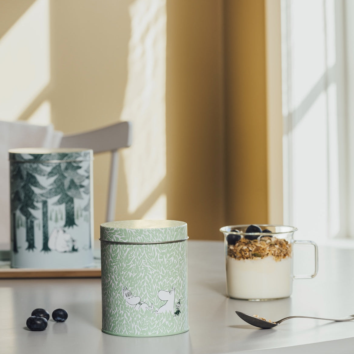 A light green cylindrical tin with a white patterned design sits on a white table next to a glass mug of yogurt and granola. In the background, another matching patterned tin and a tissue box are visible in a softly lit room with warm yellow walls and window shadows.