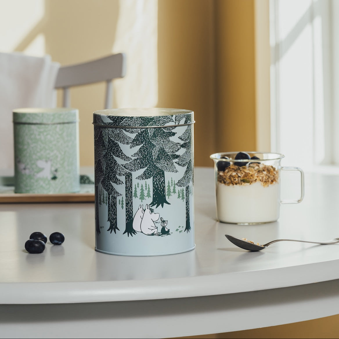 A cylindrical tin with a forest illustration stands on a white table in a bright interior. A matching green tin sits in the background on a tray, and a glass cup of yogurt with granola and blueberries is to the right.