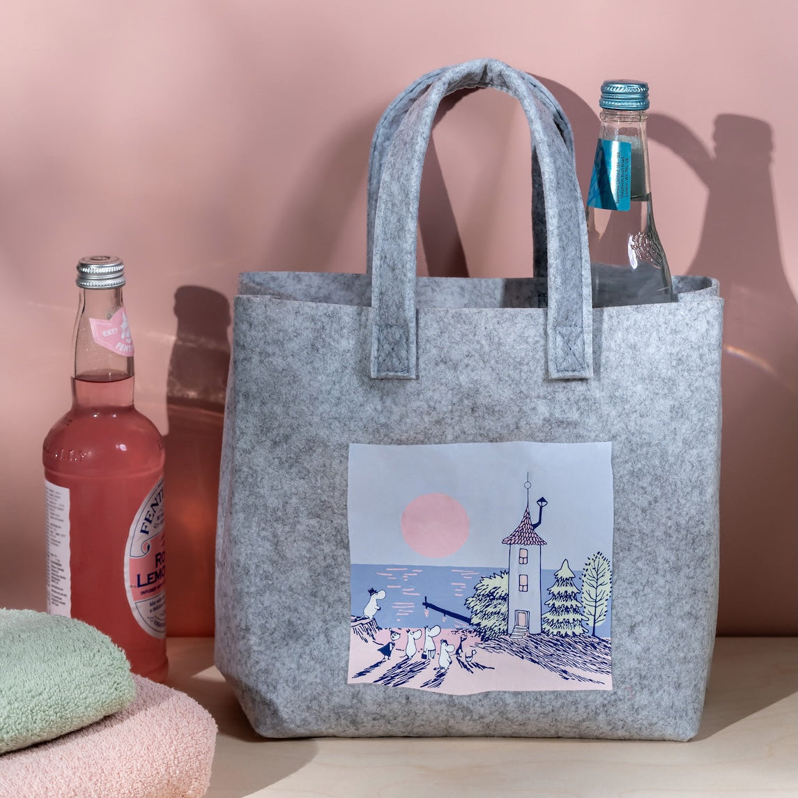 Moomin tote bag with a beach sunset scene print, placed on a pink surface with drinks and towels.