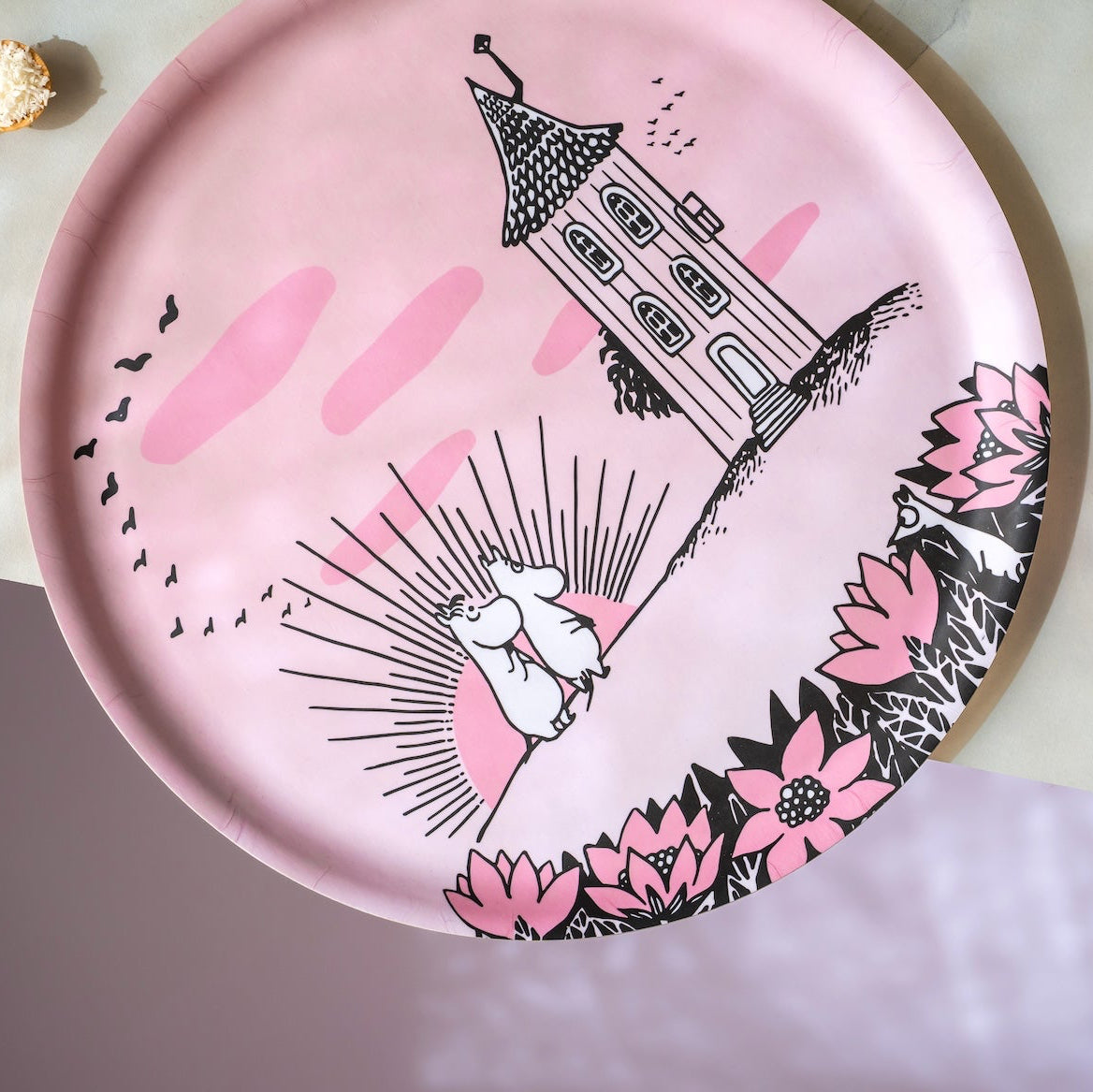 A round pink serving platter with a whimsical illustration of a rabbit on a swing, a tower, pink clouds, and floral accents, styled on a light marble surface with snacks nearby.