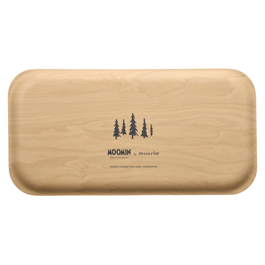 A rectangular metal tray with rounded corners, featuring a winter landscape illustration of Moomin characters and pine trees. The tray is styled on a light-colored table next to a cup of coffee, decorative pom-poms, and ribbons.