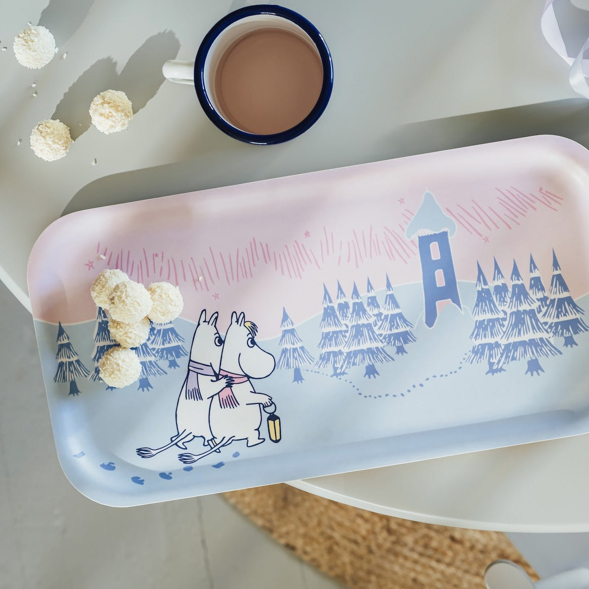 A rectangular metal tray with rounded corners, featuring a winter landscape illustration of Moomin characters and pine trees. The tray is styled on a light-colored table next to a cup of coffee, decorative pom-poms, and ribbons.