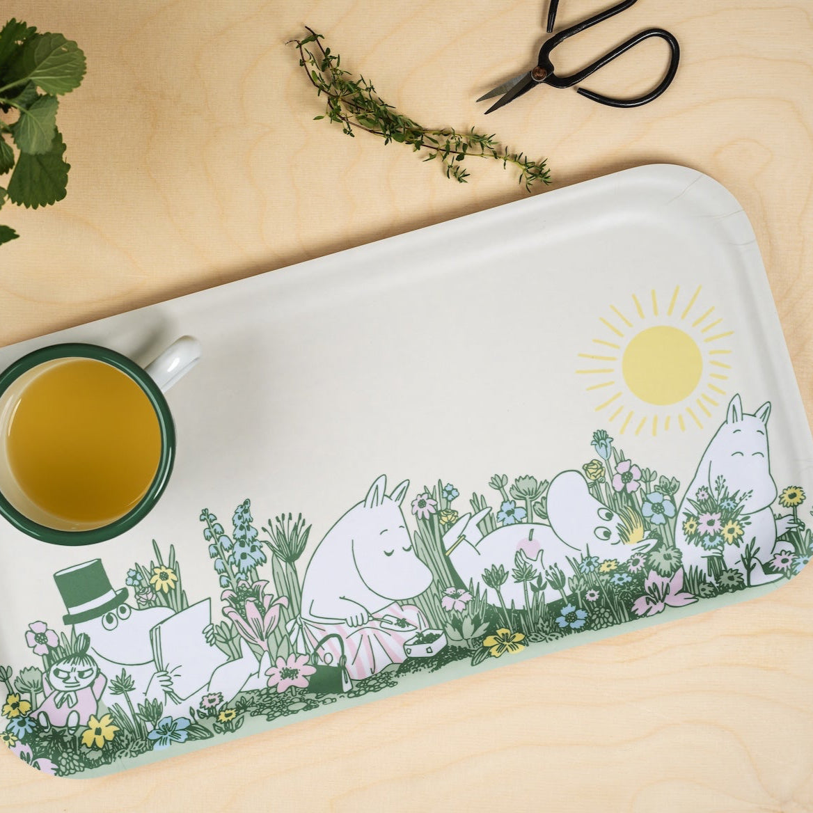 Muurla Moomin In The Garden Tray 43 x 22cm shown on a plain tabletop next to herbs and an Moomin enamel mug with a green rim 