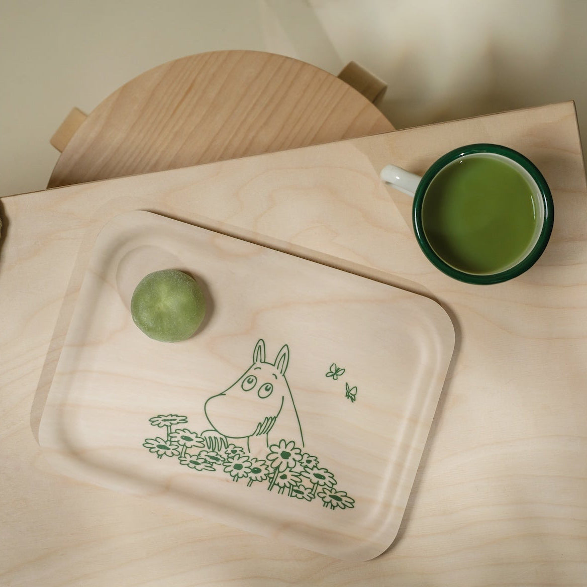 Tray with a Moomin character design on a wooden surface next to a cup of green tea.