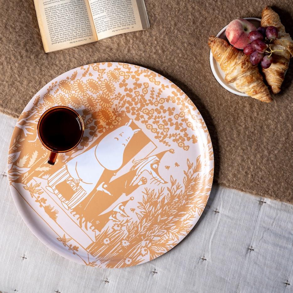 Large round Moomin Wild Garden Tray, set on the floor with a cup of coffee on it, and a plate of croissant and a book nearby.