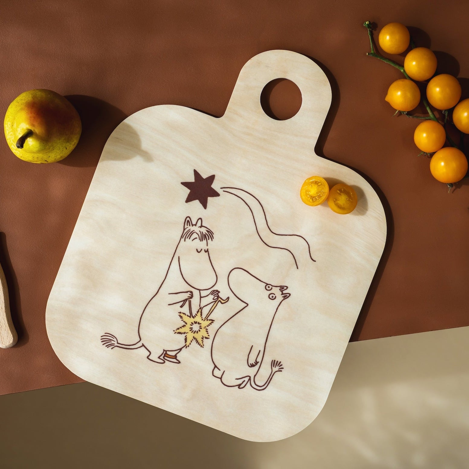 A dark brown Moomin wooden cutting board with a white print of cartoon mice and stars, styled on a light wood countertop with a knife, a pear, and a branch of yellow cherry tomatoes.