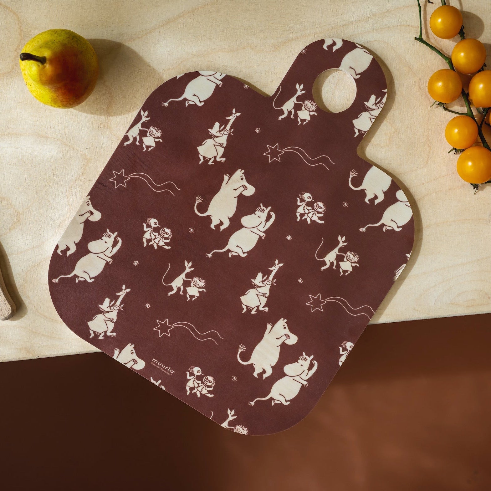 A dark brown Moomin wooden cutting board with a white print of cartoon mice and stars, styled on a light wood countertop with a knife, a pear, and a branch of yellow cherry tomatoes.