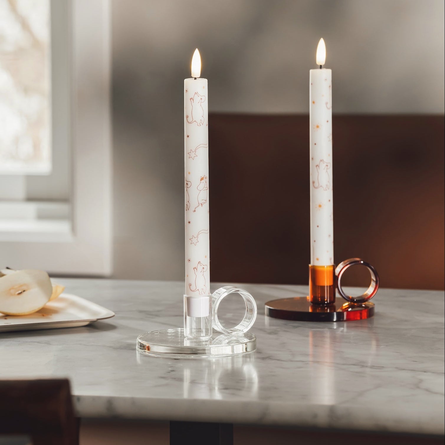A lit taper LED Moomin candle with a white base and delicate brown line illustrations stands in a dark amber glass holder with a ring-shaped loop, placed on a light wood table with a pear and chair in the background.