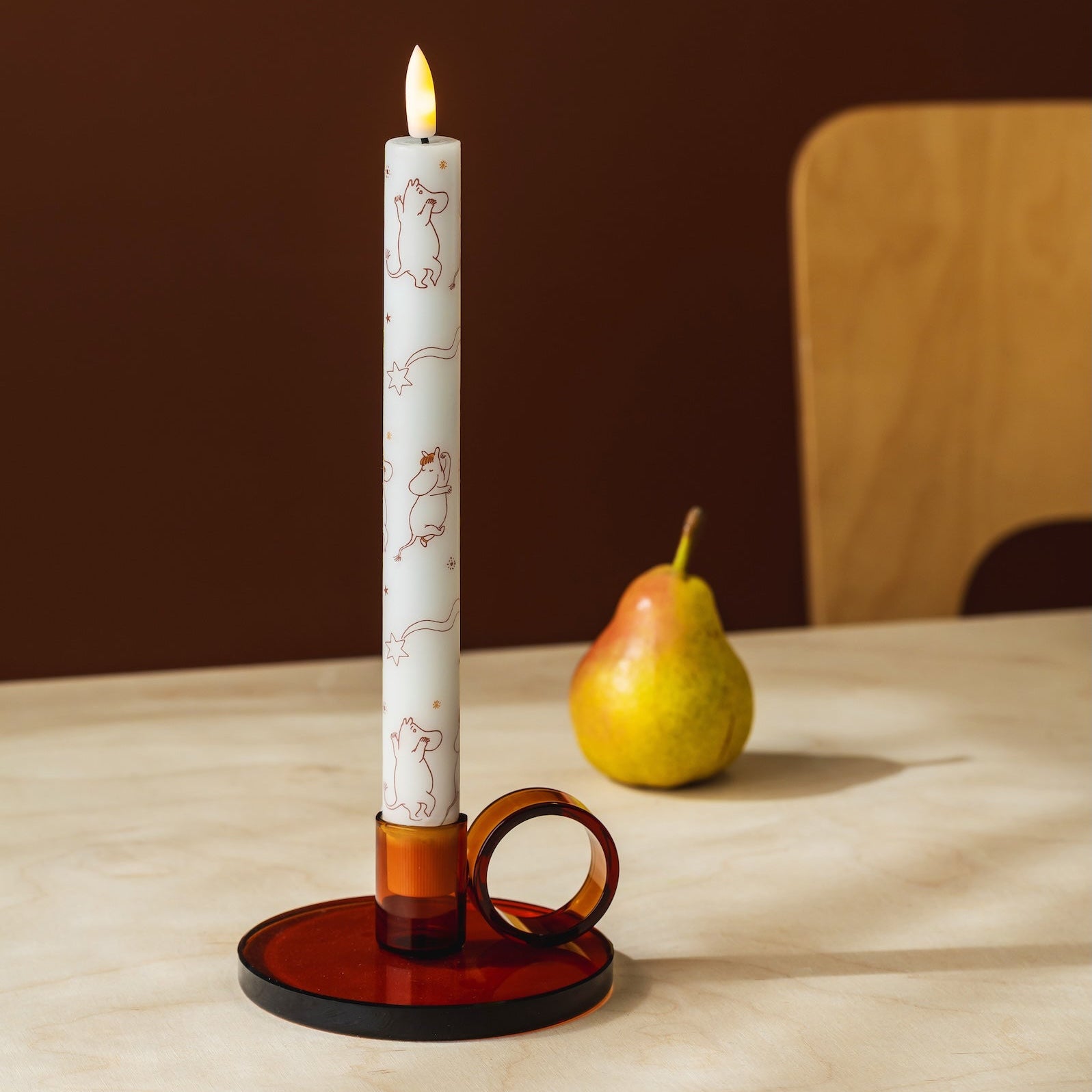 A lit taper LED Moomin candle with a white base and delicate brown line illustrations stands in a dark amber glass holder with a ring-shaped loop, placed on a light wood table with a pear and chair in the background.