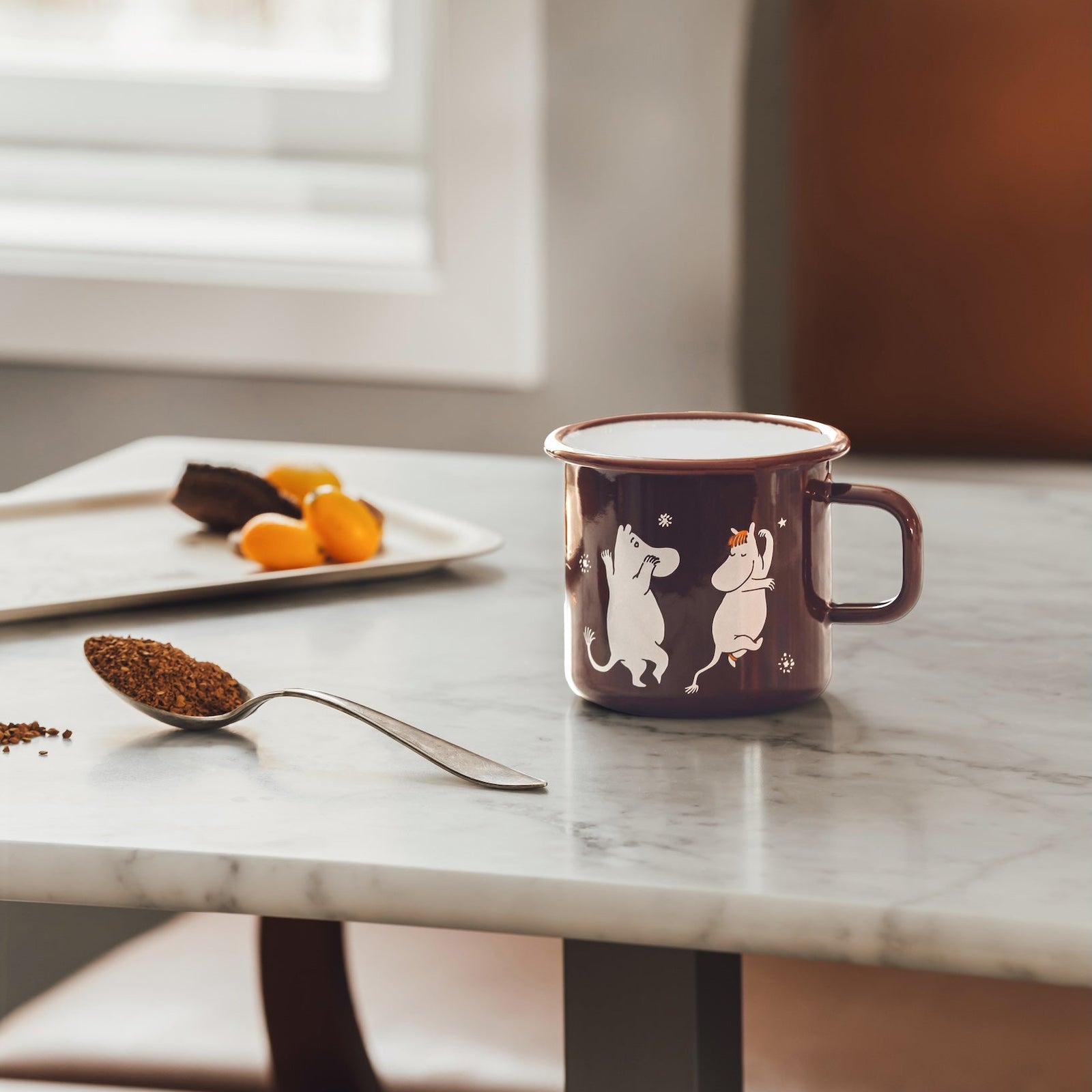A dark brown Moomin Comet enamel mug with white Moomin character illustrations sits on a light marble table in a cozy indoor setting near a window, with a spoon of coffee and a plate of fruit nearby.