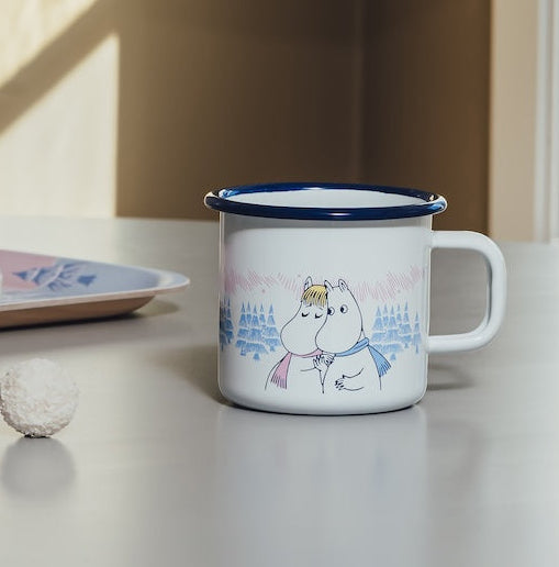 A white enamel mug with a blue rim and a cute snowman illustration, styled on a light gray table in a warm, sunlit room.