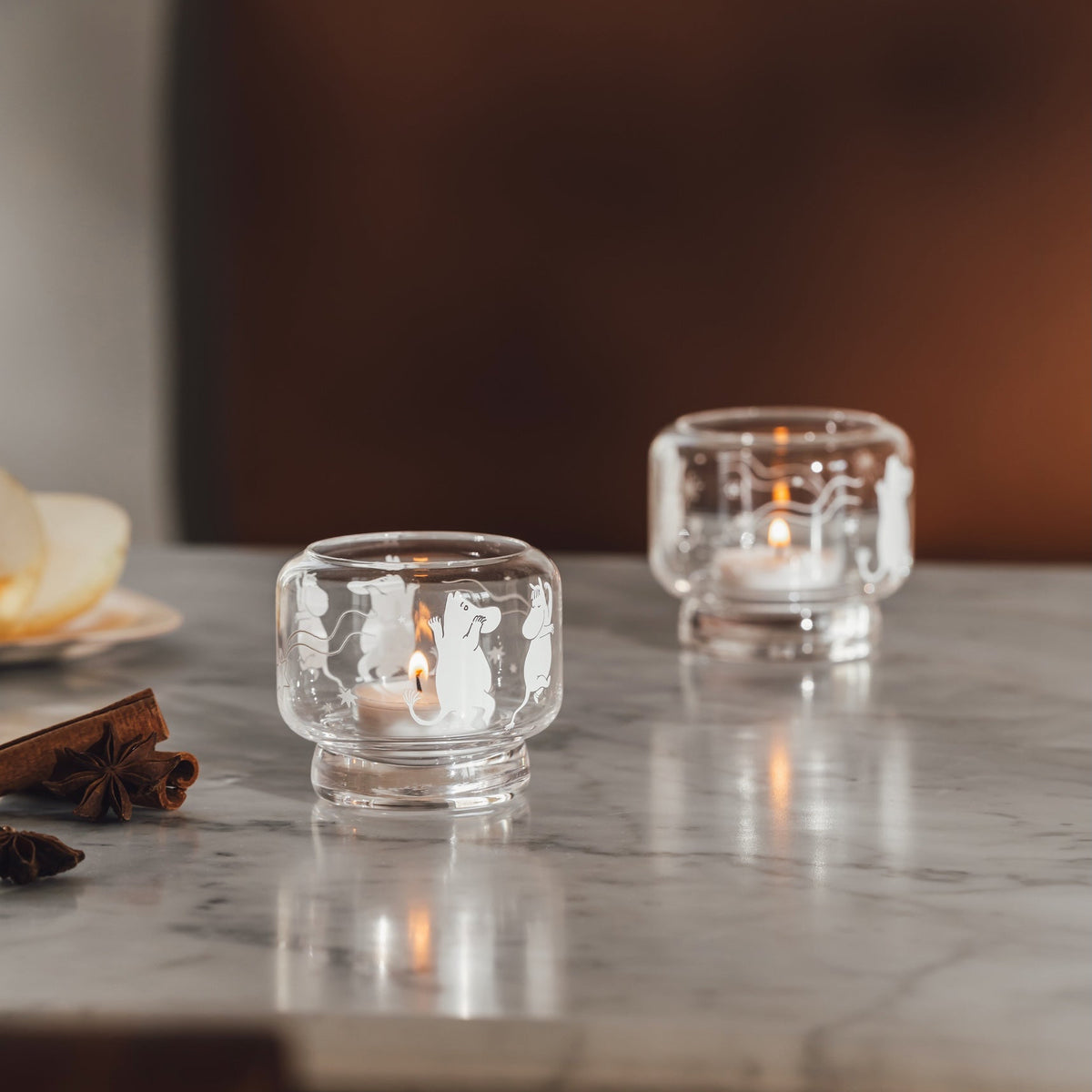 Two small clear Moomin glass candle holders with a textured, hammered finish, each containing a lit tea light candle, set on a polished gray marble tabletop with a warm brown leather chair in the background.