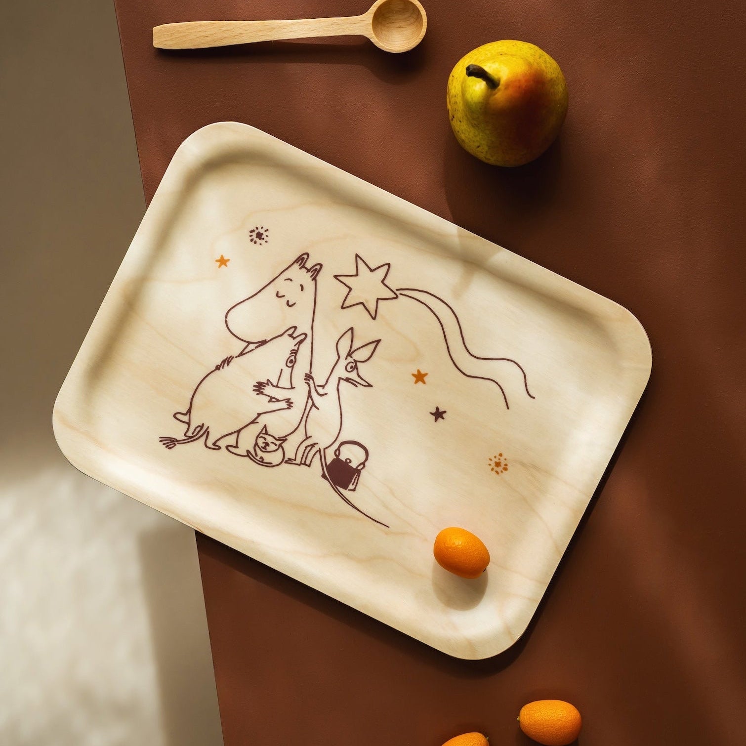 A rectangular Moomin wooden serving tray with rounded corners, featuring a hand-drawn illustration of a child and dog under stars. The tray is styled on a dark brown wooden table alongside a small wooden spoon, a pear, and three small oranges.