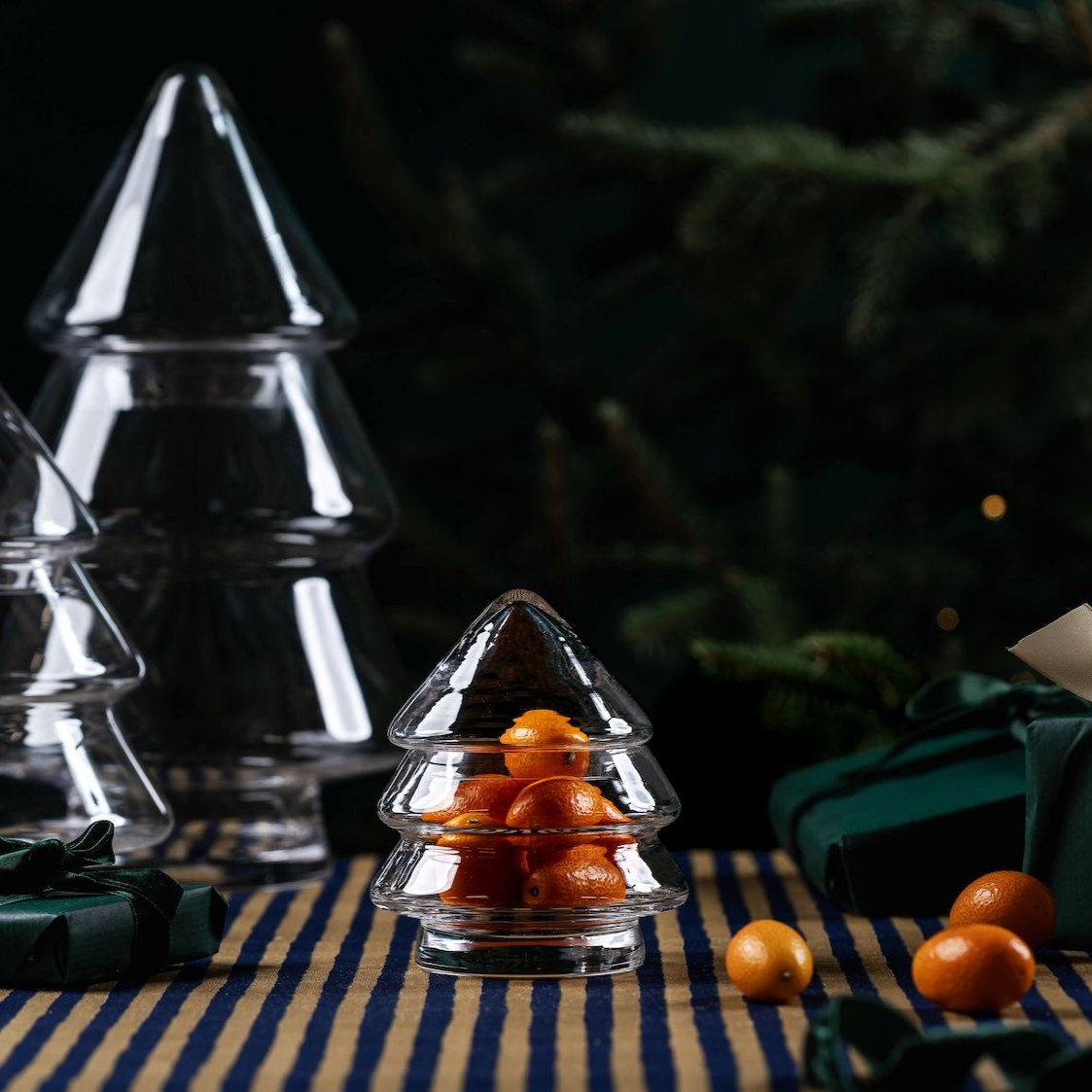 Muurla Design Glass Christmas Tree Jars in three sizes. shown on a table with a blue striped tablecloth and a Christmas tree and gifts in the background.