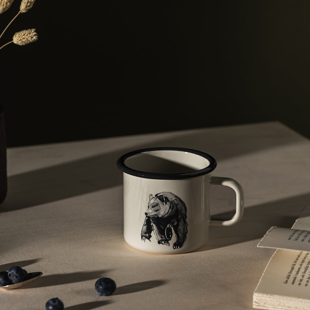 Teemu Jarvi Bear Enamel Mug by Muurla, on a table next to an open book.