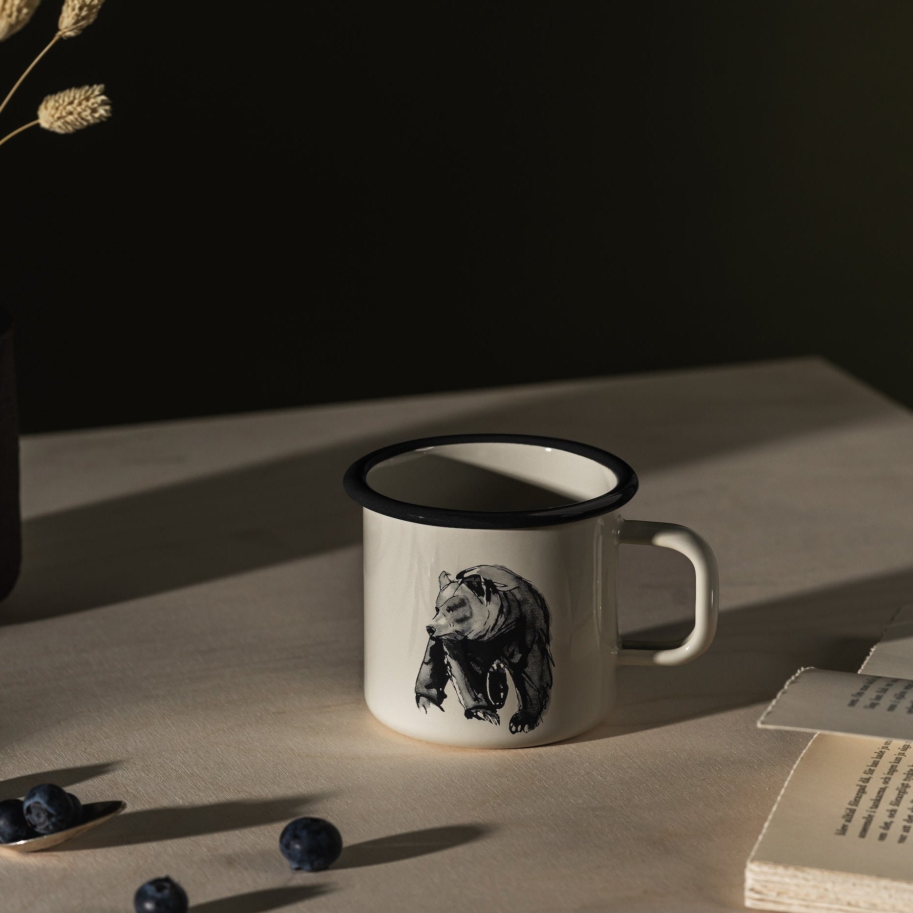 Teemu Jarvi Bear Enamel Mug by Muurla, on a table next to an open book.