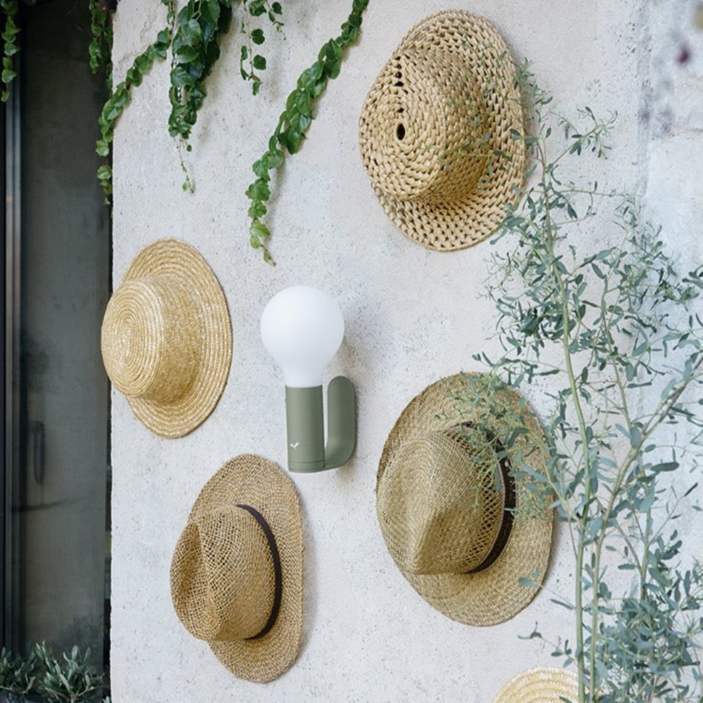 Fermob Aplô portable Lamp in Cactus attached to the wall using a Fermob wall bracket in Cactus surrounded by straw hats and wall plants.