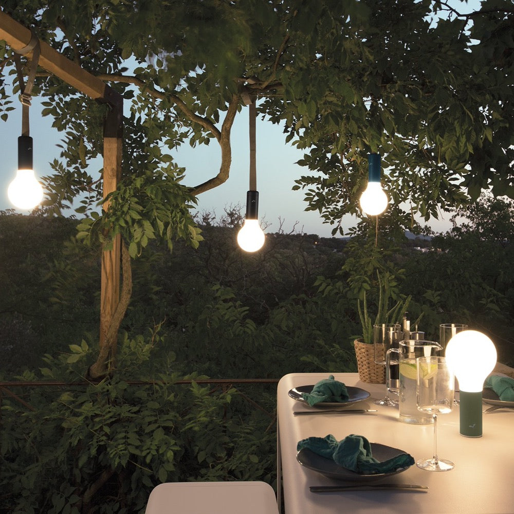 Green Garden setting lit up by four Fermob Aplô portable Lamps hanging from branches and on the table.