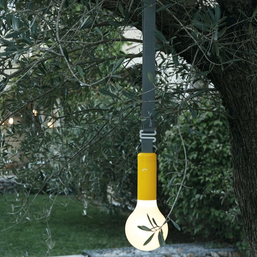 Fermob Aplô portable Lamp in Honey hanging from a tree from the Aplô suspension strap.