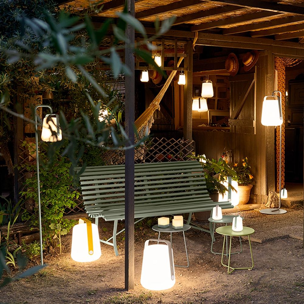 Large Fermob lighting collection glowing over a garden setting and the inside of a wooden building including a Fermob Balad Spike Stand in Cactus glowing over a bench.