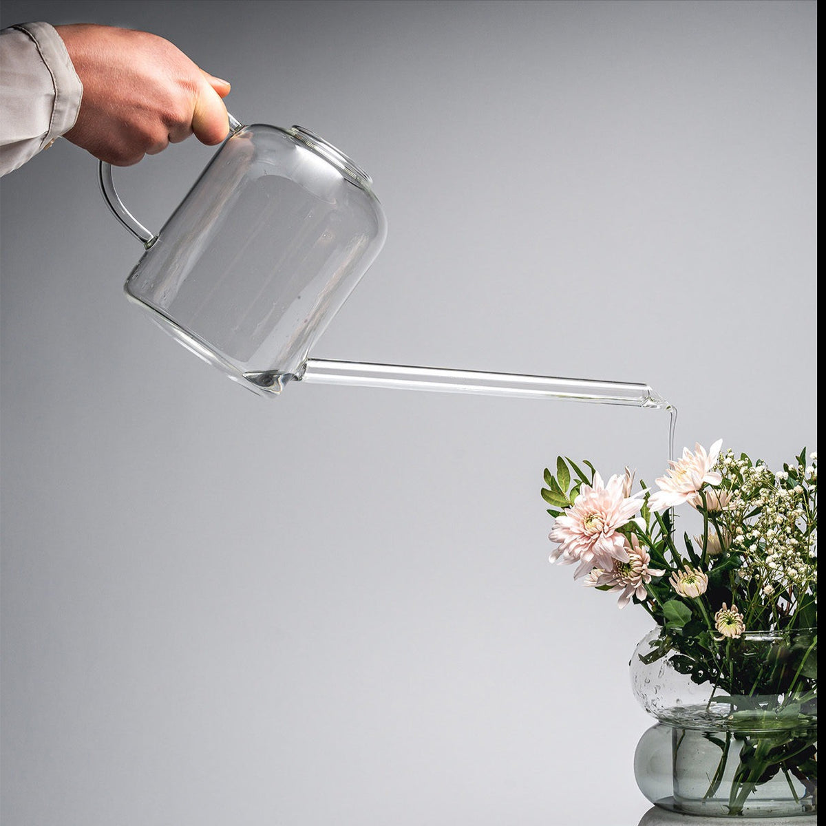 Water being poured from Muurla Design&#39;s Glass Watering Cans into a Muurla Bagel Vase full of flowers.