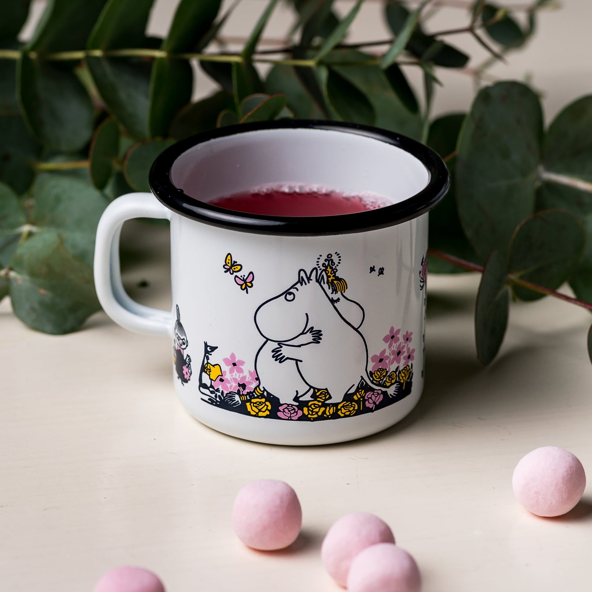 Moomin Enamel Mug, on a table next to marshmallow sweets and a plant.