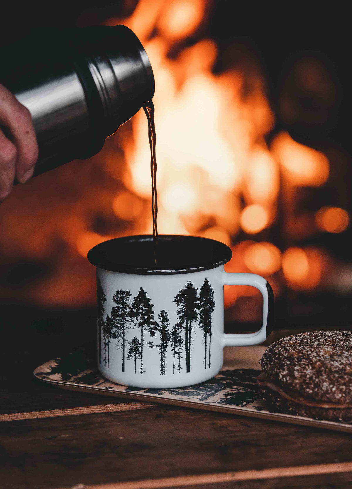 Muurla Design The Forest Enamel Mug being filled by a travel flask in front of a campfire